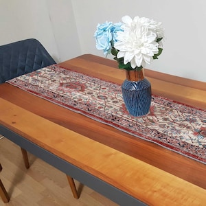 Puede incluir: Una mesa de comedor de madera con un camino de mesa estampado y un jarrón azul con flores blancas y azul claro. El camino de mesa tiene un borde rojo y diseños florales. El jarrón es azul oscuro con un patrón geométrico.