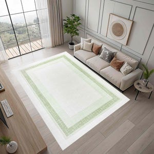 May include: A rectangular area rug with a white center and three concentric borders of varying shades of green. The rug is placed on a light wood floor in a living room setting, with a sofa and large window visible.