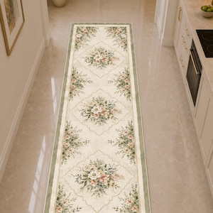 May include: A long, rectangular runner rug featuring a cream-colored background with a repeating pattern of floral bouquets in shades of pink, white, and green. The rug is framed by a sage green border and includes decorative geometric outlines.