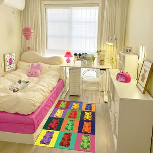 Pode incluir: Um tapete de urso de gelatina colorido com um design pop art. O tapete está em um quarto com uma cama rosa, uma escrivaninha branca e prateleiras brancas.