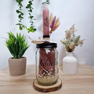 Puede incluir: Un tarro de vidrio con tapa negra y base de madera, lleno de velas rosas y papel decorativo. El tarro tiene una etiqueta que dice "Glückslichter" y un trébol de cuatro hojas. Una sola vela rosa con la palabra "Liebe" escrita en ella está colocada en la parte superior del tarro.