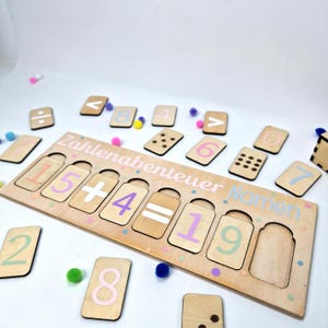 Könnte beinhalten: Pädagogisches Holzpuzzle mit dem Text "Zahlenabenteuer Namen". Das Puzzle zeigt Zahlen und mathematische Symbole in Pastellfarben mit passenden Zahlenkarten. Bunte Pompons sind um das Puzzle herum verstreut.