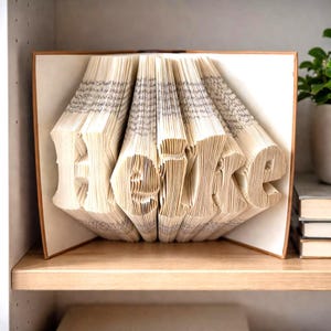 Könnte beinhalten: Eine Buchskulptur mit gefalteten Seiten, die den Namen Heike bilden. Die Seiten sind cremefarben mit sichtbarem schwarzem Text, und das Buch ist geöffnet, wobei die festen Einbände an den Seiten positioniert sind, um die Falttechnik zu zeigen.