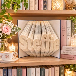 Op de afbeelding: Een gevouwen boekkunstsculptuur met de naam Steffie in het midden. De pagina's zijn nauwkeurig gevouwen om de letters te vormen, wat een driedimensionaal effect creëert. Het boek staat op een houten plank tussen andere boeken.