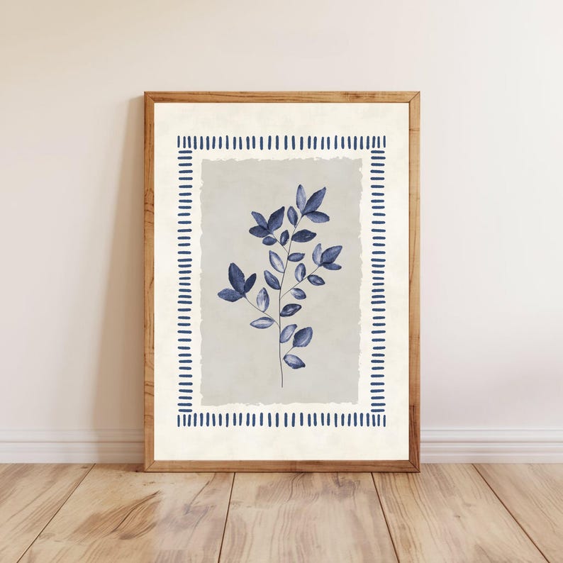 May include: A framed botanical print featuring a sprig of blue leaves against a light gray background. The artwork is bordered by a dashed blue line on a cream-colored background, encased in a wooden frame. The print is displayed on a wooden floor.