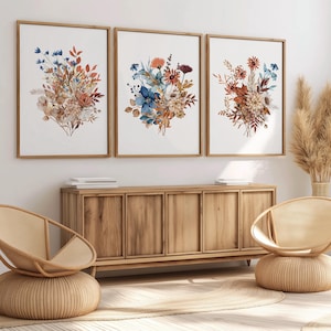 Könnte beinhalten: Drei gerahmte Drucke von Aquarell-Blumensträußen mit blauen, braunen und weißen Blumen. Die Drucke hängen an einer weißen Wand über einem Holzschränk.