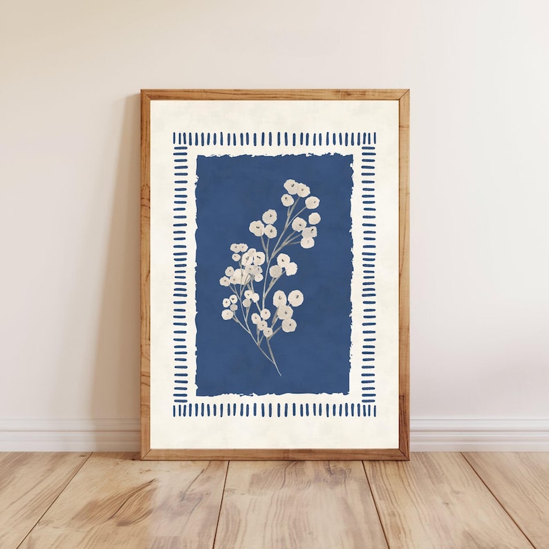May include: A framed botanical print featuring a white floral arrangement against a deep blue background. The artwork is enclosed within a cream-colored border with vertical line accents and a natural wood frame. The print is displayed on a wooden floor.
