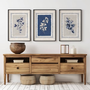 May include: Three framed botanical prints with black frames. Each print features a different floral arrangement in shades of blue and white against a neutral background. The prints are displayed above a wooden cabinet with a large vase and decorative items.