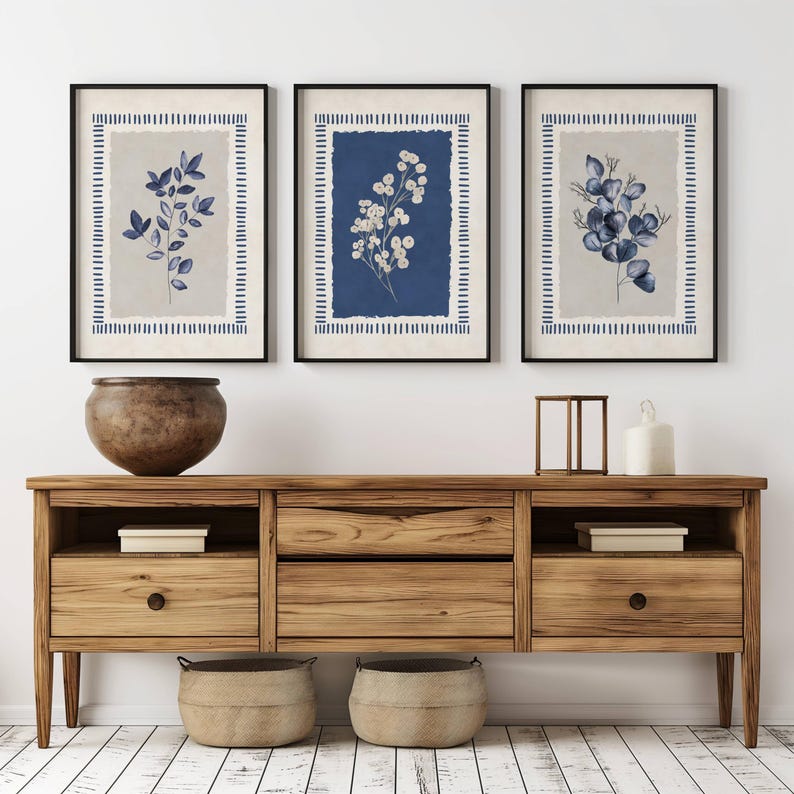 May include: Three framed botanical prints with black frames. Each print features a different floral arrangement in shades of blue and white against a neutral background. The prints are displayed above a wooden cabinet with a large vase and decorative items.