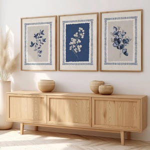 May include: Three framed botanical prints with blue and white floral designs. The prints are in wooden frames and hang above a wooden cabinet. A tall white vase with dried pampas grass and decorative wooden bowls complete the decor.