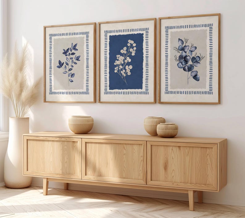 May include: Three framed botanical prints with blue and white floral designs. The prints are in wooden frames and hang above a wooden cabinet. A tall white vase with dried pampas grass and decorative wooden bowls complete the decor.
