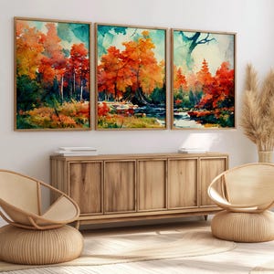 May include: Three framed watercolour paintings depicting a forest scene with a stream running through it. The trees are in shades of orange, red, and yellow, suggesting autumn. The paintings are hung on a white wall above a wooden cabinet.