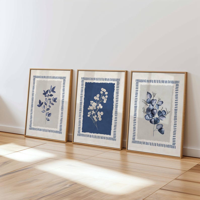 May include: Three framed botanical prints. Each print features a different blue and white floral design on a neutral background. The frames are a light wood color, and the prints are leaning against a white wall.