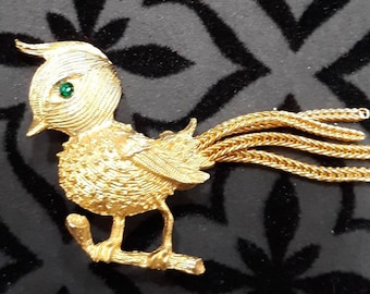 Può includere: Una spilla a forma di uccello dorata con un occhio di strass verde e una lunga coda a catena.