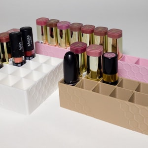 May include: Three rectangular lipstick organizers with a honeycomb pattern on the exterior sides. One organizer is white, one is light pink, and one is tan. Each unit features multiple square compartments for storing lipsticks in various colors.