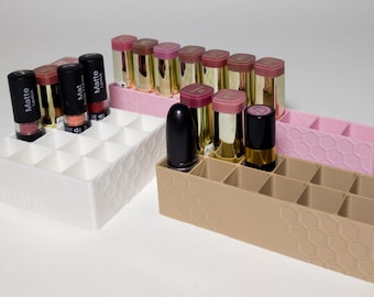 May include: Three rectangular lipstick organizers with a honeycomb pattern on the exterior sides. One organizer is white, one is light pink, and one is tan. Each unit features multiple square compartments for storing lipsticks in various colors.