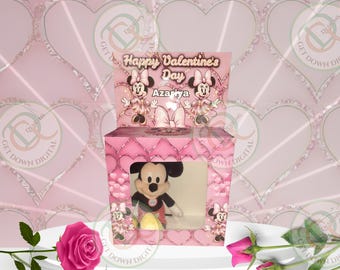 May include: Pink gift box with a clear window showing a plush Mickey Mouse toy inside. The box has a pink and white heart pattern and says "Happy Valentine's Day Azariya" on the top.