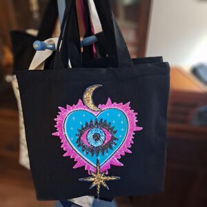 Könnte beinhalten: Schwarze Tragetasche mit einem herzförmigen Design mit Auge, Mond und Stern. Das Herz ist türkis mit einem rosa Rand und einem goldenen Halbmond. Das Auge ist blau mit schwarzen Details, und der Stern ist golden.