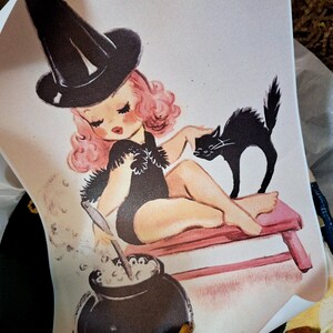 May include: A retro illustration on a white backdrop depicts a witch with pink hair and a black hat. She sits on a pink bench, stroking a black cat. A cauldron bubbles with a potion, creating a Halloween theme.