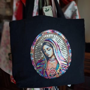 May include: A black tote bag featuring a large oval patch of the Virgin Mary. The image is surrounded by a border of iridescent, multi-colored sequins. The central figure wears a head covering and a garment adorned with a single pink rose graphic.