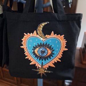 May include: Black tote bag with a heart-shaped design featuring a blue eye, orange sunburst, gold crescent moon, and star. The bag has black handles and is made of a sturdy fabric. The design is embellished with sequins and beads.