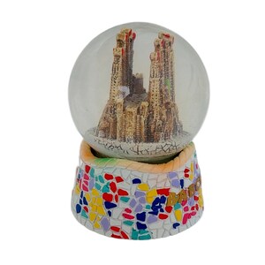 May include: A glass snow globe with a miniature replica of the Sagrada Familia church in Barcelona, Spain. The base of the snow globe is decorated with colorful mosaic tiles and the text "Barcelona" in gold lettering.