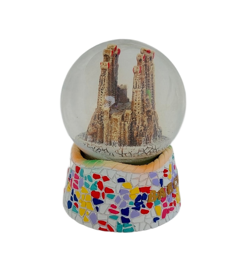 May include: A glass snow globe with a miniature replica of the Sagrada Familia church in Barcelona, Spain. The base of the snow globe is decorated with colorful mosaic tiles and the text "Barcelona" in gold lettering.