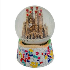 May include: A glass snow globe with a miniature replica of the Sagrada Familia church in Barcelona, Spain. The base of the snow globe is decorated with a colorful mosaic pattern.