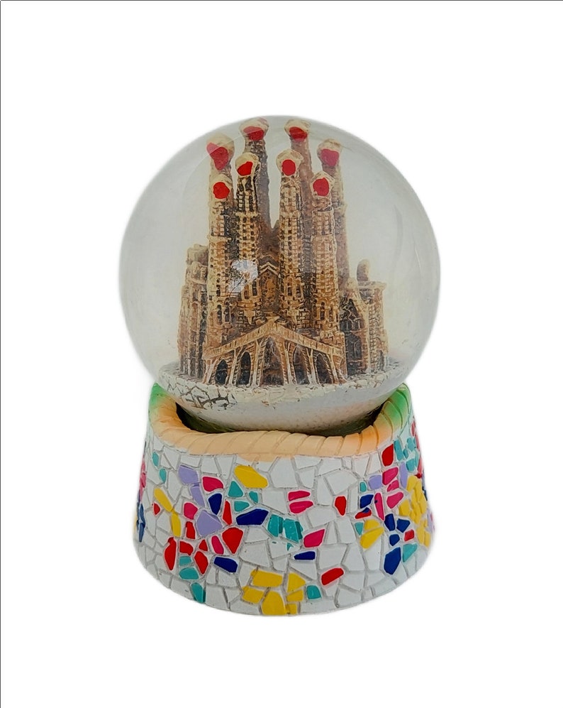 May include: A glass snow globe with a miniature replica of the Sagrada Familia church in Barcelona, Spain. The base of the snow globe is decorated with a colorful mosaic pattern.