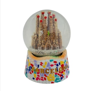 May include: A glass snow globe with a miniature replica of the Sagrada Familia church in Barcelona, Spain. The base of the snow globe is decorated with colorful mosaic tiles and the word "BARCELONA" in gold lettering.