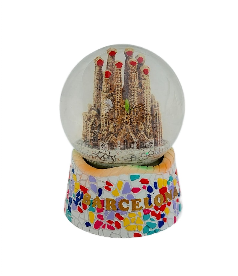 May include: A glass snow globe with a miniature replica of the Sagrada Familia church in Barcelona, Spain. The base of the snow globe is decorated with colorful mosaic tiles and the word "BARCELONA" in gold lettering.