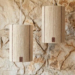 Puede incluir: Dos lámparas colgantes cilíndricas con material beige similar a una cuerda envuelto alrededor de las pantallas. Líneas verticales marrones acentúan los lados. Pequeñas etiquetas cuadradas marrones están adheridas a la parte inferior de cada pantalla. Las luces cuelgan de cables blancos contra una pared de piedra.