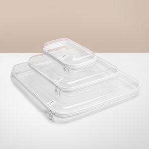 May include: Three clear plastic packing cubes with white zips and white trim. The cubes are nested inside each other, with the largest cube on the bottom and the smallest cube on top. The cubes are labelled "C-Gear" on the front.