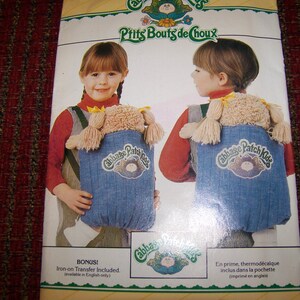 May include: A vintage Cabbage Patch Kids backpack pattern. The pattern features a child wearing the backpack, which is denim blue with the Cabbage Patch Kids logo. The pattern also includes the text "Pits Bouts de Choux".