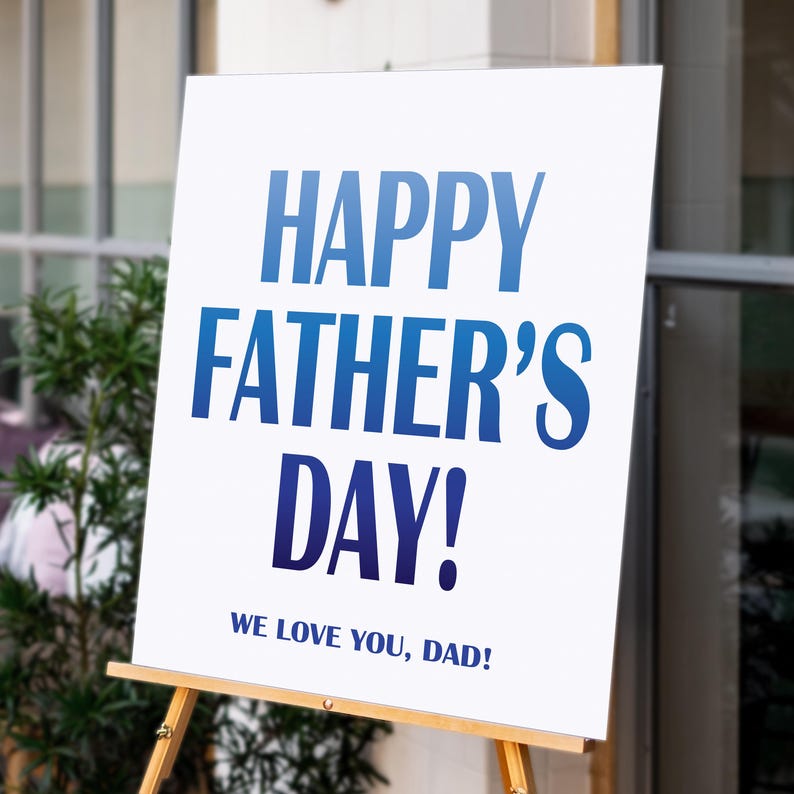Op de afbeelding: Een wit bord op een houten ezel met de tekst "HAPPY FATHER'S DAY!" in blauw. Daaronder staat "WE LOVE YOU, DAD!". Het bord staat buiten, met groen en een gebouw op de achtergrond.