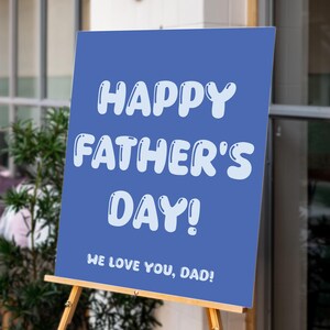 Op de afbeelding: Een blauw bord met de tekst "HAPPY FATHER'S DAY!" in witte bubbelletters. Hieronder staat "WE LOVE YOU, DAD!". Het bord wordt weergegeven op een houten ezel, met groen en een gebouw op de achtergrond.