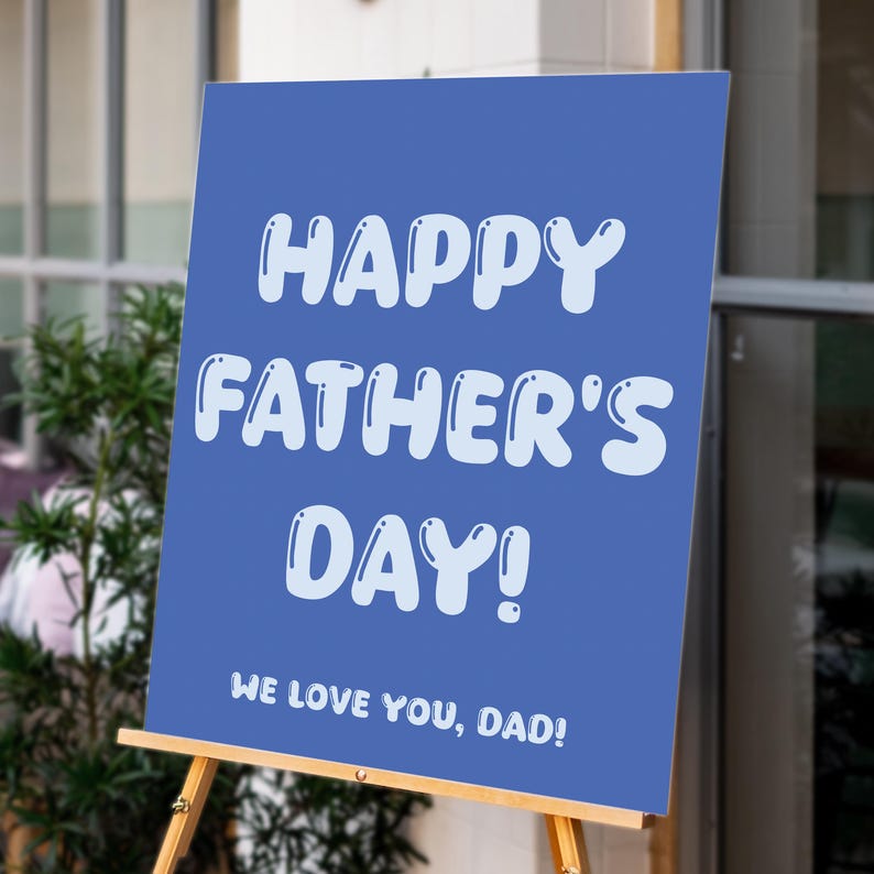 Op de afbeelding: Een blauw bord met de tekst "HAPPY FATHER'S DAY!" in witte bubbelletters. Hieronder staat "WE LOVE YOU, DAD!". Het bord wordt weergegeven op een houten ezel, met groen en een gebouw op de achtergrond.