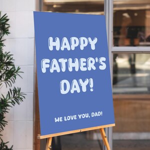 Op de afbeelding: Een blauw bord op een houten ezel met de tekst "HAPPY FATHER'S DAY!" in witte, bubbelachtige letters. Hieronder staat op het bord "WE LOVE YOU, DAD!". Het bord wordt buiten een gebouw tentoongesteld.