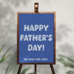Op de afbeelding: Een houten ezel met een blauw bord met de tekst "HAPPY FATHER'S DAY!" in witte bubbelletters. Daaronder staat "WE LOVE YOU, DAD!". Het bord is omlijst met bruin hout.