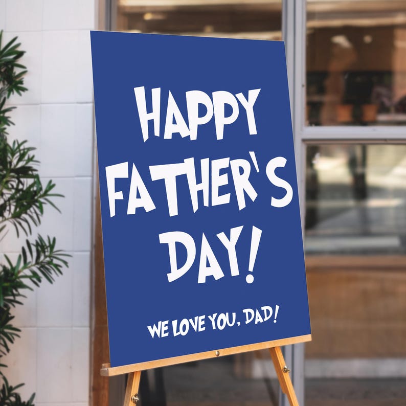 Op de afbeelding: Een blauw bord met witte tekst met de tekst "HAPPY FATHER'S DAY!" en "WE LOVE YOU, DAD!". Het bord staat op een houten ezel, perfect om Vaderdag te vieren.