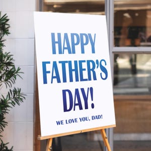 Op de afbeelding: Een wit bord op een houten ezel met de tekst "HAPPY FATHER'S DAY!" in blauw. Onder de hoofdtekst staat de zin "WE LOVE YOU, DAD!" in een kleiner lettertype. Het bord staat buiten.