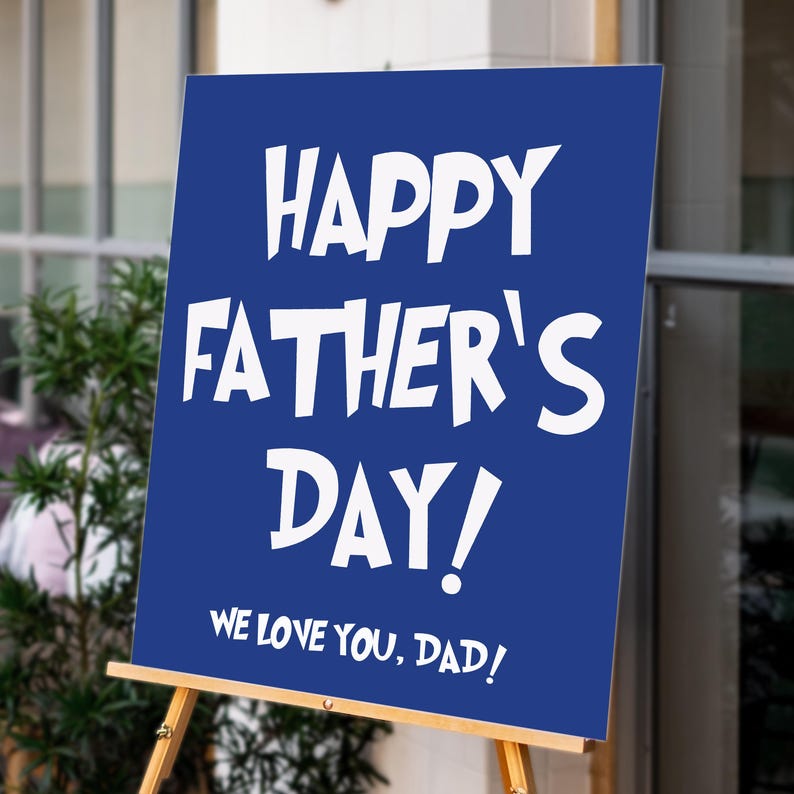 Op de afbeelding: Een blauw bord op een houten ezel met de tekst "HAPPY FATHER'S DAY! WE LOVE YOU, DAD!" in witte letters. Het bord wordt buiten weergegeven, met groen op de achtergrond.
