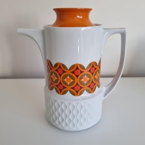 Könnte beinhalten: Weiße Keramik-Kaffeekanne mit orangefarbenem Deckel und Griff. Die Kanne hat ein dekoratives Band aus orangefarbenen, braunen und gelben geometrischen Formen. Der untere Teil hat ein strukturiertes, wellenförmiges Design.