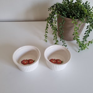 Op de afbeelding: Twee witte keramische ramekins met een geribbelde buitenkant en een tomatenontwerp in het midden. De ramekins staan op een wit oppervlak, met een potplant op de achtergrond. Ideaal voor het serveren van individuele porties.