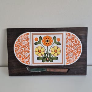 Puede incluir: Una tabla de quesos rectangular de madera marrón oscura con tres azulejos de cerámica incrustados. El azulejo central muestra un motivo floral con flores naranjas y amarillas, mientras que los dos laterales presentan volutas naranjas y blancas. Un cuchillo de queso dentado con mango de madera está fijado debajo de los azulejos.