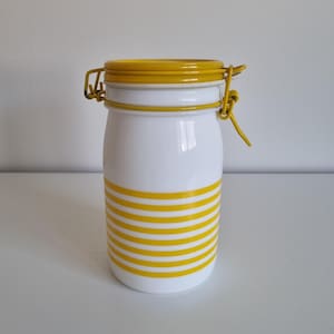 Op de afbeelding: Witte keramische pot met een geel deksel en gele strepen. De pot heeft een metalen sluiting.