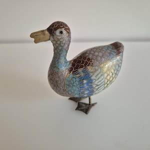 Può includere: Statuetta decorativa a forma di anatra con un design cloisonné dettagliato. L'anatra presenta una testa marrone, un becco giallo e un corpo ornato da motivi blu, viola e marroni. La statuetta ha zampe in metallo ed è ambientata su uno sfondo bianco.