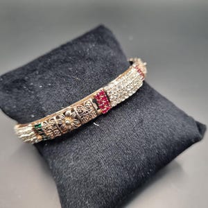 Könnte beinhalten: Goldfarbenes Armreif-Armband mit aufwendigen floralen und geometrischen Mustern. Das Armband ist mit klaren, roten und grünen Edelsteinen besetzt. Es liegt auf einem schwarzen Samtkissen.