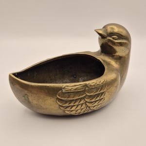 Könnte beinhalten: Eine messingfarbene, vogelförmige Schale mit detailliertem Flügeldesign. Die Schale hat einen tiefen, offenen Innenraum und einen kleinen, spitzen Schnabel. Der Vogel hat eine warme, goldene Farbe, und die Schale wird wahrscheinlich zu dekorativen Zwecken verwendet.