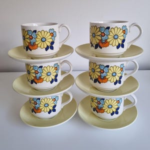 Può includere: Set di sei tazze da tè e piattini vintage con un motivo floreale in tonalità di giallo, blu, arancione e verde. Le tazze e i piattini hanno uno sfondo bianco con un bordo giallo.